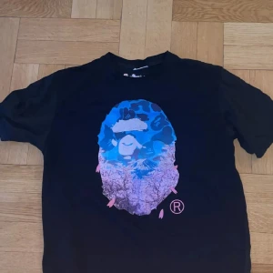 Svart Bape t-shirt med blått tryck - Svart t-shirt från Bape med ikoniskt ape-huvud i blått och lila tryck framtill. Klassisk passform och rund hals. Perfekt för dig som gillar streetwear och vill sticka ut med en unik design.