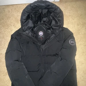 Svart jacka från Canada Goose - Säljer en svart dunjacka från Canada Goose med klassisk patch på ärmen och stor huva. Jackan är fylld med dun och har detaljer i äkta päls. Perfekt för kalla vinterdagar och har en stilren, tidlös look. Men dragkedjan är sönder o fick den i present så tror bara att den är äkta 