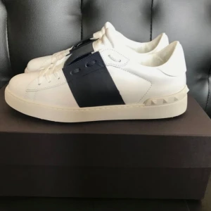 Valentino Garavani sneakers vit/blå - Snygga vita sneakers från Valentino Garavani med bred mörkblå rem över snörningen och ikoniska nitar på hälen. Skorna är i skinn och har en stilren, modern design som passar perfekt till jeans eller dressade outfits.