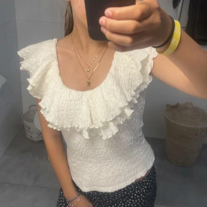 Vit volangblus med smock - Supersöt vit blus med smockad kropp och stora volangdetaljer vid axlar och halsringning. Ärmlös modell som ger en romantisk vibe, perfekt till sommaren. Toppens struktur är lätt rynkad och materialet känns mjukt och luftigt.