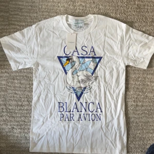 Vit t-shirt Casablanca med svanmotiv - Casablanca t-shirt i vitt med stort tryck av en svan och blå triangeldesign framtill. Klassisk rund hals och korta ärmar. Snygg och clean look med texten 'Casa Blanca Par Avion' i blått. Perfekt för dig som gillar streetstyle och unika prints.