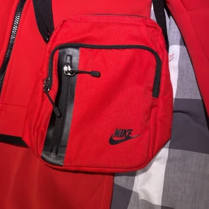 Röd axelväska från Nike - Säljer en snygg röd axelväska från Nike med svart logga och detaljer. Väskan har justerbar rem, flera fack med dragkedja och är gjord i slitstark polyester. Perfekt storlek för mobilen, plånboken och andra smågrejer. Enkel att matcha till streetwear.