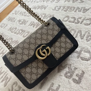 Gucci GG Supreme axelväska med kedja - Säljer en ikonisk Gucci axelväska i GG Supreme-mönster med svarta skinnkanter och guldfärgad kedja. Väskan har ett stort GG-spänne framtill och justerbar axelrem i kedja med skinnparti för extra komfort. Perfekt för dig som vill ha en klassisk och trendig look.