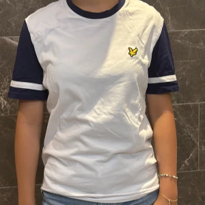 Vit och marinblå t-shirt Lyle & Scott - Vit t-shirt från Lyle & Scott med marinblå ärmar och krage. Klassisk gul logga på bröstet. Ärmarna har vit randdetalj och t-shirten är i mjuk bomull. Perfekt för en chill och sportig look.