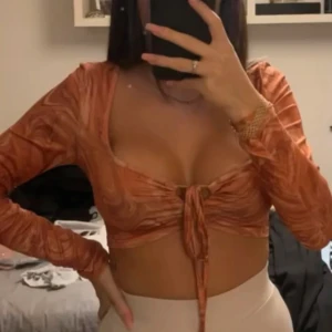 Croppad omlottopp i orange mönster - Trendig croppad omlottopp i orange med swirl-mönster. Toppen har långa ärmar, djup ringning och snygg knytning framtill. Perfekt för dig som vill sticka ut med en cool och färgstark look.