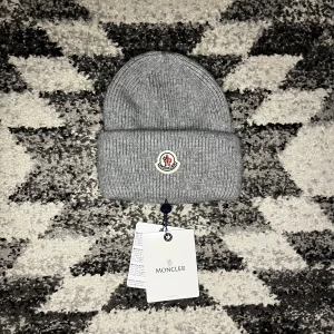 Moncler mössa - Snygg ljusgrå ribbstickad mössa från Moncler med klassisk uppvikt kant och ikonisk logotyp framtill. Perfekt accessoar för kyliga dagar och ger en clean look till din outfit.
