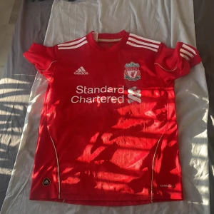 Liverpool FC röd Adidas matchtröja - Liverpool FCs officiella matchtröja från Adidas i rött med vita detaljer och klubbmärke broderat på bröstet. Tröjan har korta ärmar, rund hals och är tillverkad i lätt funktionsmaterial (Climacool) som andas. Klassisk design med vita ränder på axlarna och sponsortryck framtill. Från säsongen 2010/2011