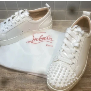 Vita Christian Louboutin sneakers med nitar - Snygga vita sneakers från Christian Louboutin med ikoniska röda sulor och coola vita nitar på tån. Skorna har snörning och är tillverkade i skinn för en lyxig känsla. Perfekta för dig som vill sticka ut med exklusiv streetstyle.