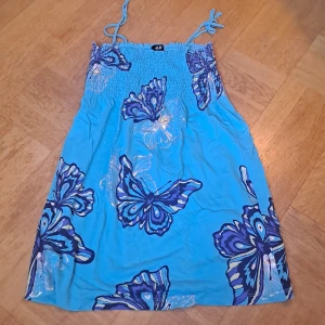 Turkos sommarklänning med fjärilar  - Supersöt turkos klänning från H&M med stora fjärilar i blå och lila toner samt silvriga detaljer. Klänningen har smock upptill och tunna axelband som knyts. Barnstorlek 170, men mycket stretch, så kan passar xs-m. Kan eventuellt även användas till kjol. 🩷 Färg stämmer bäst överens med sista bilden. 🩷 Fanns någon fläck men ska gå bort i  tvätt. 🩷