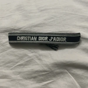 Christian Dior J'Adior armband svart - Snyggt armband från Christian Dior med texten 'J'Adior' broderad i vitt på svart bakgrund. Armbandet har knytning och små tofsar i ändarna, vilket ger en cool och trendig look. Perfekt accessoar för att lyfta din outfit.