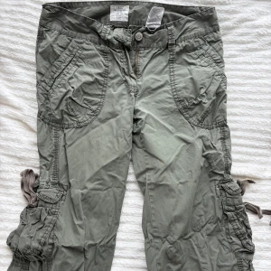 Gröna cargopants med fickor - Säljer ett par gröna cargopants med stora fickor på sidorna och bak, samt snörningsdetaljer vid benen. Byxorna har avslappnad passform och är tillverkade i bomull, perfekta för en chill och streetig stil.
