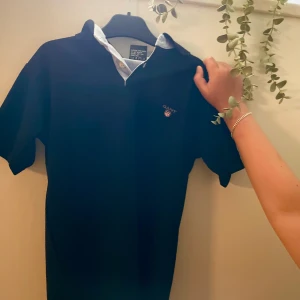 Svart pikétröja från GANT - Svart pikétröja från GANT med klassisk krage och ljusblå insida vid knäppningen. Tröjan har korta ärmar, två knappar framtill och GANT-logga broderad på bröstet. Perfekt för en clean och stilren look.