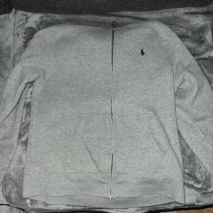 Grå hoodie från Polo Ralph Lauren - Snygg grå hoodie från Polo Ralph Lauren med dragkedja framtill och klassisk blå broderad logga på bröstet. Tröjan har huva, känguruficka och är tillverkad i mjuk bomullsmix. Perfekt för chill dagar eller när du vill ha en clean look.