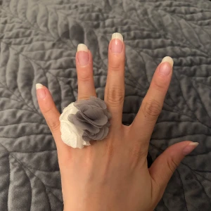 Stor ring med tygblommor i grått och vitt - Unik statementring med två stora tygblommor i grått och vitt. Blommorna är fluffiga och sitter på en silverfärgad ringbas. Storlek justerbar. Perfekt för dig som vill sticka ut och addera en cool detalj till din look.