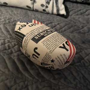 Glasögonfodral med tidningsmönster - Snyggt glasögonfodral med tidningsinspirerat mönster och amerikanska flaggor. Fodralet är hårt och skyddar dina glasögon på ett stilrent sätt. Färgerna är främst vit, svart och röd med inslag av blått. Perfekt för dig som vill ha något unikt i väskan.