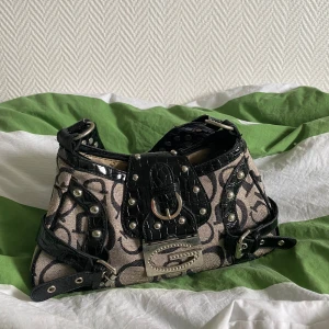 Y2k väska🥳 - Cool axelväska med beige och svart logomönster, detaljer i svart glansig skinnimitation och silverfärgade nitar. Väskan har ett stort G-spänne framtill, bältesdetaljer och bred axelrem. Perfekt statement piece för dig som gillar att sticka ut.🥳 Använt men Fint skick överlag men bandets sidor är lite synliga (se bild 5) har sina skavanker