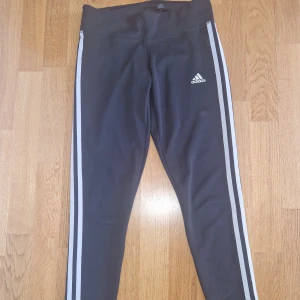 Svarta Adidas leggings med ränder - Svarta leggings från Adidas med klassiska vita ränder längs sidorna och Adidas-logga på låret. Modellen är tight och högmidjad, tillverkad i stretchigt Climalite-material som andas. Perfekta för träning eller chill dagar.