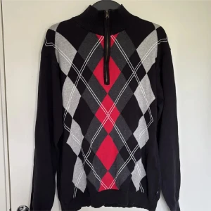 Argyletröja med half zip från Trend - Snygg långärmad tröja från Trend i storlek XL med klassiskt argylemönster i svart, grått och rött. Tröjan har hög krage och half zip-dragkedja framtill. Perfekt för dig som gillar preppy stil och vill sticka ut lite extra.