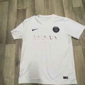Vit PSG x Balmain fotbollströja Nike - Snygg vit fotbollströja från Nike i samarbete med PSG och Balmain. Tröjan har korta ärmar, rund hals och tryck med Balmain Paris på bröstet samt PSG-logga och Nike swoosh. Tillverkad i lätt och bekvämt material som passar perfekt för fotboll eller streetwear.