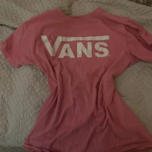 Rosa Vans t-shirt med logga - Säljer en rosa t-shirt från Vans i storlek S med stor vit logga framtill. Klassisk rund halsringning och korta ärmar. Tillverkad i 100% bomull för en skön och avslappnad känsla. Perfekt för dig som gillar streetwear och vill sticka ut med färg. Det är dock två små hål på framsidan som man dock inte märker på men kan synas om man kollar nog.
