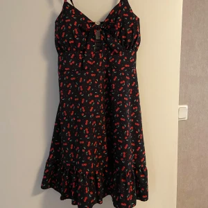  Känning med körsbär  - Söt svart kortklänning med röda körsbärsmönster, smala axelband och en rosett framtill. Klänningen har volang nertill och är perfekt för sommardagar.