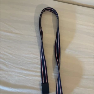 Randigt marinblått tygbälte - Snyggt tygbälte med marinblå bas och ränder i vitt och rött. Bältet har en enkel svart spänne och passar perfekt till jeans eller shorts. Stilren och sportig look med klassiska färger och randigt mönster.