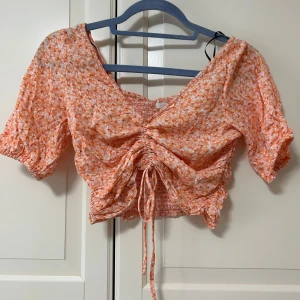 Blommig croppad blus med snörning - Superfin croppad blus i orange och vitt med blommigt mönster. Toppen har korta puffärmar, smockad rygg och snygg snörning framtill som ger en cool rynkad effekt. Man kan även dra ner resåren och få toppen längre!
