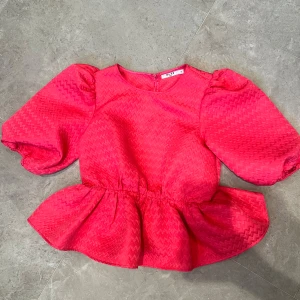 Rosa volangblus från NA-KD - Superfin rosa blus från NA-KD med puffiga korta ärmar och volang nertill. Blusen har ett subtilt geometriskt mönster i tyget och rund halsringning. Perfekt för dig som vill sticka ut med en färgstark och trendig look.