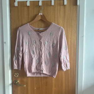 Rosa kofta med paljetter från Noa Noa - Supersöt ljusrosa kofta från Noa Noa med glittriga paljettdetaljer över hela tröjan. Koftan har knappar framtill, trekvartsärm och är stickad i ett mjukt material. Perfekt för dig som gillar en feminin och lekfull stil!