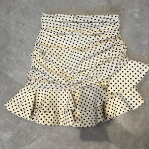 Prickig volangkjol från Zara - Supersöt kort kjol från Zara i beige med svarta prickar. Kjolen har volangkant nertill och snygga rynkade detaljer på sidorna. Perfekt för dig som gillar lekfulla och trendiga plagg med lite extra attityd.
