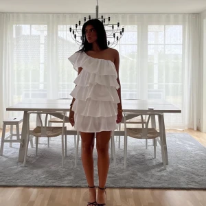 Vit studentklänning Anielle  - Säljer en superfin vit kortklänning med flera lager volanger och oneshoulder-design. Perfekt för dig som vill sticka ut med en romantisk och trendig look. Klänningen har en luftig passform och är riktigt snygg till sommarens alla tillfällen.