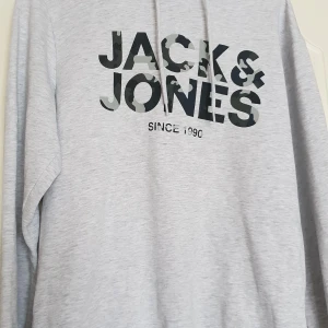 Ljusgrå hoodie från Jack & Jones - Ljusgrå hoodie med tryck i svart och camouflage på bröstet där det står 'Jack & Jones'. Tröjan har huva med dragsko och är tillverkad i mjukt material. Perfekt för en avslappnad stil.