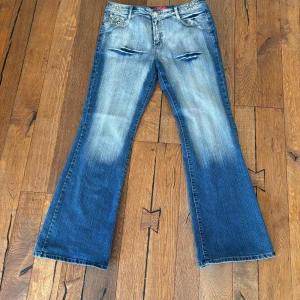 Blå bootcut jeans med slitningar - Säljer ett par blå bootcut jeans med slitningar framtill och ljus tvätt. Jeansen är lågmidjade och har  broderade detaljer på bakfickorna. Jag säljer dessa jeans då de är stora för mig och har därför ingen användning. På etiken står det strl. 44, men skulle påstå att det passar för strl. 42.                                 Frakt ingår ej ❤️