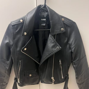 Svart skinnjacka från Pull&Bear - Säljer en svart skinnjacka från Pull&Bear med klassisk bikerstil. Jackan har silvriga dragkedjor, knappar och bälte i midjan. Snygg krage och fickor framtill. Perfekt för dig som vill ha en edgy look. Pris kan såklart diskuteras. Ställ jättegärna frågor om du har några funderingar. 