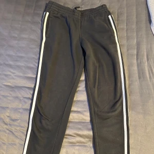 Svarta träningsbyxor med vita ränder från Adidas - Svarta mjukisbyxor från Adidas med vita ränder längs sidorna och logga på vänster ben. Byxorna har resår i midjan och vid bensluten, samt en sportig och bekväm passform. De är oanvända.