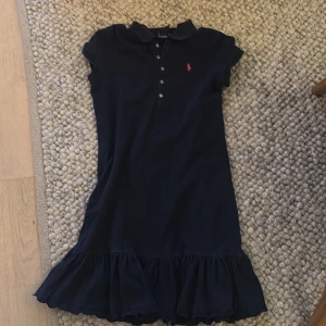 Mörkblå pikéklänning med volang från Ralph Lauren - Supersöt mörkblå pikéklänning från Ralph Lauren med liten krage, knappar framtill och volang nertill. Klassisk logga broderad på bröstet och fina broderade detaljer längs volangkanten. Storlek 12/14