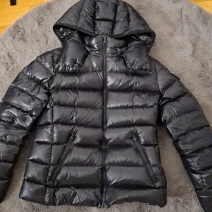 Svart dunjacka från Moncler med huva - Säljer en svart, glansig dunjacka från Moncler med huva och dragkedja framtill. Jackan har två snedställda fickor med dragkedja och en ärmficka med Moncler-logga. Perfekt för kyliga dagar. Den är köpt i butik. 