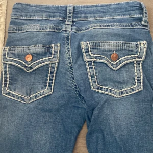 bootcut jeans - Säljer ett par blå jeans med bootcut passform och snygga kontrastsömmar på bakfickorna. Jeansen har dekorativa knappar på fickorna och klassisk femficksdesign. Perfekta för dig som gillar en retroinspirerad look.