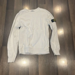 Vit sweatshirt från Stone Island Junior - Säljer en vit långärmad sweatshirt från Stone Island Junior med klassisk logotyp-patch på ärmen. Tröjan har rund halsringning och ribbade muddar vid ärmslut och nederkant. Perfekt för en clean och stilren look.