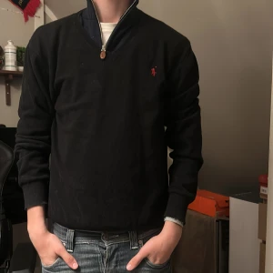 Svart långärmad tröja från Ralph Lauren - Svart långärmad tröja från Ralph Lauren med liten röd logga broderad på bröstet. Tröjan har hög krage med dragkedja och ribbade muddar vid ärmslut och nederkant. Perfekt för en stilren och avslappnad look.