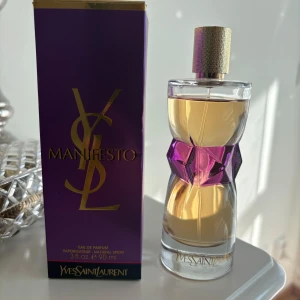 Manifesto Eau de Parfum från Yves Saint Laurent - En lyxig parfymflaska från Yves Saint Laurent, Manifesto, med en elegant design i glas och en lila detalj på mitten. Doften är förpackad i en lila och guldig kartong med stora YSL-bokstäver. Flaskan rymmer 90 ml.