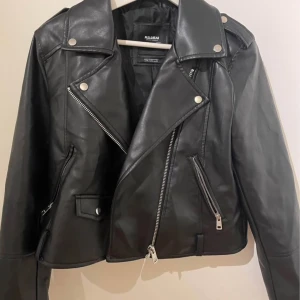 Svart skinnjacka från Pull&Bear - Säljer en svart skinnjacka från Pull&Bear i storlek XL. Jackan har klassisk bikerstil med sned dragkedja, silverfärgade detaljer och flera fickor med dragkedjor. Perfekt för dig som vill ha en cool och tidlös look.