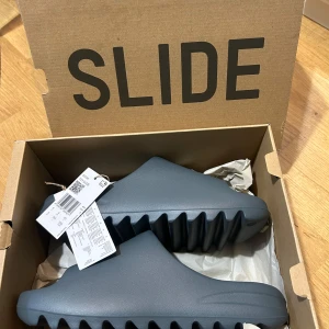 Adidas Yeezy Slide grå - Säljer ett par gråa Adidas Yeezy Slide med chunky sula och öppen tå. Skorna är tillverkade i syntetmaterial och har en modern, minimalistisk design. Perfekta för dig som gillar streetwear och vill ha något bekvämt och stilrent.