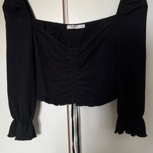 Svart croppad topp från NA-KD - Trendig svart croppad topp från NA-KD med långa puffärmar och volangdetaljer vid ärmslut. Toppen har knytning framtill och en lätt rynkad effekt på bröstet. Perfekt för att skapa en snygg och stilren look.