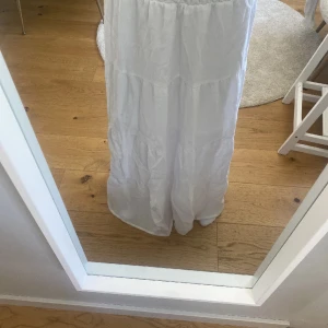 Vit långkjol från H&M - Säljer en vit långkjol från H&M med smock upptill och volangliknande sömmar längs hela klänningen. Perfekt för varma dagar och har en luftig passform. Tillverkad i ett lätt tyg som känns svalt. 