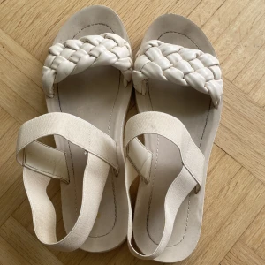 Beige sandaler med flätad rem - Säljer ett par snygga beige sandaler med bred, flätad rem över tårna och elastiskt band runt vristen. Skorna har platt sula och är perfekta för varma dagar. Passar dig som gillar stilrena och trendiga detaljer.