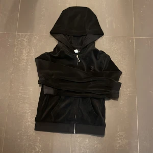 Svart velour hoodie från Juicy Couture - Säljer en svart hoodie i mjuk velour från Juicy Couture. Tröjan har dragkedja, huva, fickor framtill och ett broderat JC-logo på bröstet. Perfekt för en avslappnad och trendig look. Den är i bra skick och är inte så använd. Priset går att diskutera 🤩