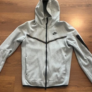 Ljusgrå hoodiejacka från Nike - Säljer en ljusgrå hoodiejacka från Nike med svart dragkedja och logga på bröstet. Jackan har huva, svarta detaljer och en ficka med dragkedja på ena ärmen. Perfekt för en sportig stil. 