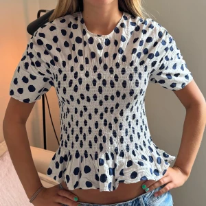 Vit blus med marinblå prickar och volang - Supersöt vit blus med marinblå prickar och smockad midja. Blusen har korta ärmar och en volangkant nedtill som ger en feminin touch. Perfekt till jeans eller kjol för en trendig look.