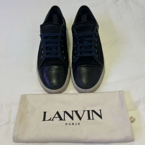 Lanvin - Säljer ett par svarta sneakers från Lanvin med vita sulor och mörkblå snörning. Skorna har en klassisk rund tå och är tillverkade i skinn. Perfekta för dig som gillar stilrena och exklusiva sneakers.